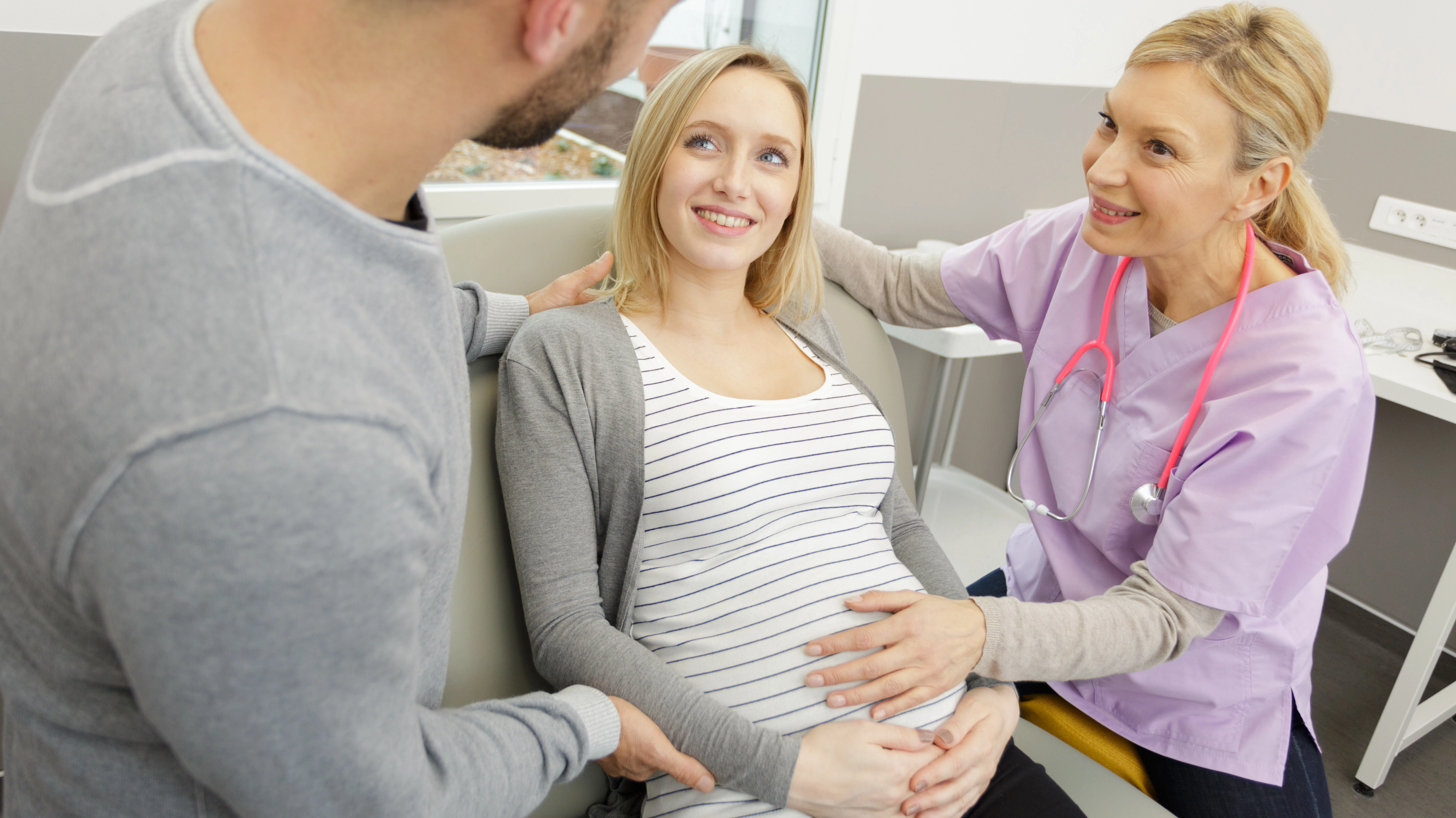 Eine Hebamme fasst auf den Bauch einer Schwangeren. Beide schauen lächelnd zur männlichen Begleitung.